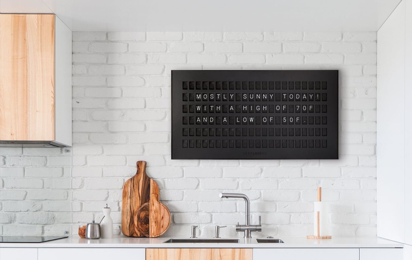 A Vestaboard split-flap display mounted on a kitchen wall showing a weather message